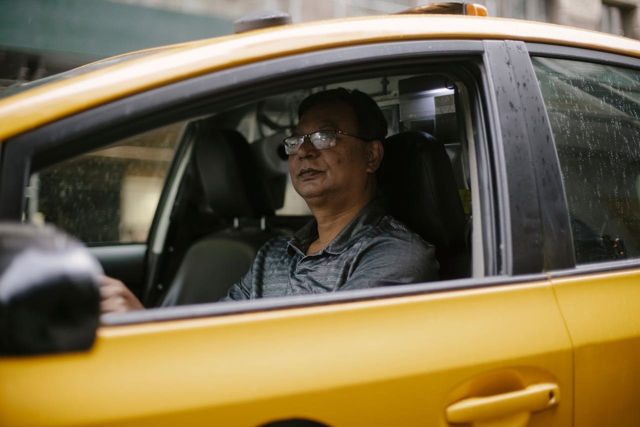 Services Middle-aged man driving a yellow taxi through a rainy cityscape.